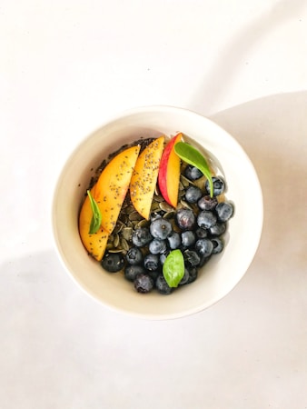 A bowl filled with a healthy mixture featuring slices of peach, fresh blueberries, chia seeds, and pumpkin seeds, garnished with a couple of green leaves. The bowl is set against a light background, creating a fresh and appetizing presentation.