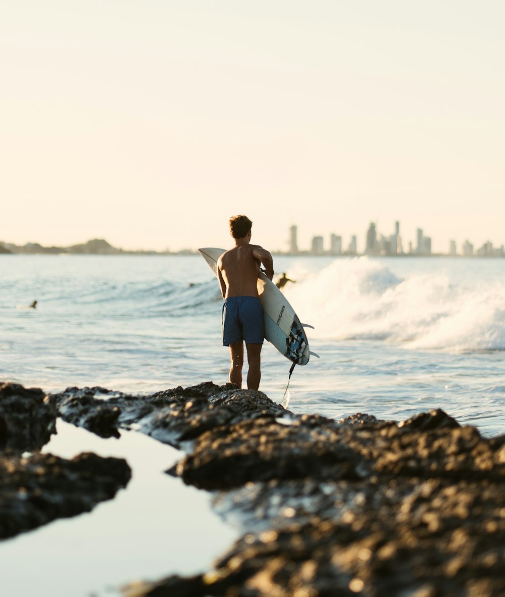 낮 동안 바다 근처의 바위에 서 있는 흰색 서핑보드를 들고 있는 파란색 비키니를 입은 여자