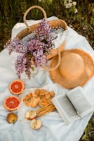 A serene picnic setup featuring a woven basket with lilac flowers, a straw hat, and a variety of snacks including croissants, crackers, sliced apples, and grapefruit. An open book lies beside the food on a white blanket, all set amidst grass and greenery.