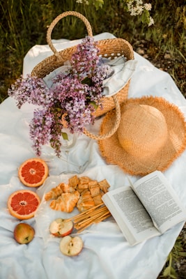 A serene picnic setup featuring a woven basket with lilac flowers, a straw hat, and a variety of snacks including croissants, crackers, sliced apples, and grapefruit. An open book lies beside the food on a white blanket, all set amidst grass and greenery.