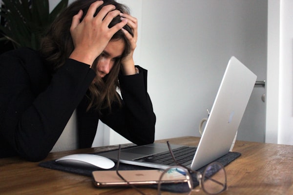 A woman holds her head in her hands, with a laptop and phone in front of her on the desk