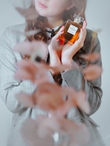 A person holds a bottle of Chanel No. 5 perfume, with hands elegantly close to their face. The individual wears a checkered blazer and has long, wavy hair. Soft-focus floral elements appear in the foreground.