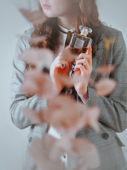 A person with long, wavy hair holds a glass bottle of Chanel perfume close to their chest. The individual is dressed in a plaid blazer and a white shirt. The foreground is blurred with a soft focus on the leaves or petals, adding an artistic touch.