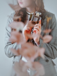 A person with long, wavy hair holds a glass bottle of Chanel perfume close to their chest. The individual is dressed in a plaid blazer and a white shirt. The foreground is blurred with a soft focus on the leaves or petals, adding an artistic touch.