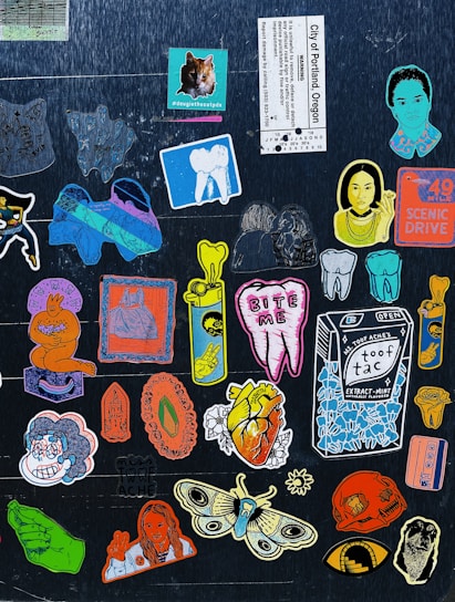 A collection of colorful, artistic stickers are arranged on a dark background. The stickers feature a variety of designs including a cat, teeth, a carton labeled 'Toof Tac', artistic portraits, a heart with the text 'Tooth Ache', and a large moth. There is a mix of cartoonish and realistic elements, with some stickers displaying text and images of hands showing peace signs.