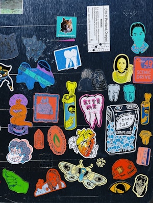 A collection of colorful, artistic stickers are arranged on a dark background. The stickers feature a variety of designs including a cat, teeth, a carton labeled 'Toof Tac', artistic portraits, a heart with the text 'Tooth Ache', and a large moth. There is a mix of cartoonish and realistic elements, with some stickers displaying text and images of hands showing peace signs.