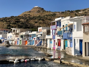 A picturesque coastal village with colorful houses built along the edge of a calm sea. The houses feature bright doors and shutters in shades of blue, red, and green, with balconies overlooking the water. Above the village, a hill covered in vegetation rises, giving the setting a peaceful and scenic backdrop.