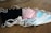 A collection of disposable face masks in different colors, including black, pink, and blue, placed on a wooden surface. The masks have white elastic ear loops.