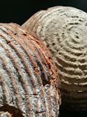 Two loaves of bread with a crusty texture and circular scoring patterns. The surface has a mix of flour dusting and a golden-brown baked appearance, with visible ridges and a rustic look.