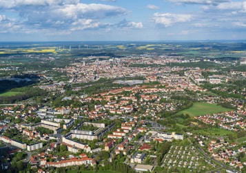 An expansive aerial view of a city showcasing a mix of residential, commercial, and industrial areas. The scene features numerous buildings with red and white roofs, sprawling green spaces, parks, and neatly arranged streets. In the distance, fields and wind turbines can be seen under a partly cloudy sky.