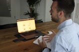 A person sits at a wooden table engaging in a conversation while gesturing with their hands. A laptop is open in front of them displaying a webpage, accompanied by a smartphone and a mouse on the table. Notebook and glasses are also nearby, with a plant in the background, creating a casual work environment.