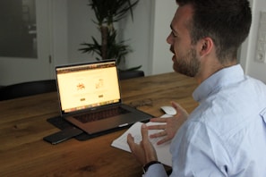 A person sits at a wooden table engaging in a conversation while gesturing with their hands. A laptop is open in front of them displaying a webpage, accompanied by a smartphone and a mouse on the table. Notebook and glasses are also nearby, with a plant in the background, creating a casual work environment.