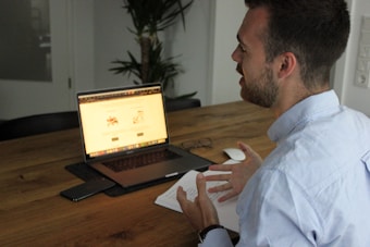 A person sits at a wooden table engaging in a conversation while gesturing with their hands. A laptop is open in front of them displaying a webpage, accompanied by a smartphone and a mouse on the table. Notebook and glasses are also nearby, with a plant in the background, creating a casual work environment.