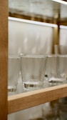A row of textured glass tumblers is displayed on a wooden and glass shelf. The glasses have a diamond or honeycomb pattern and appear to be empty. The shelf is well-lit, highlighting the clarity and design of the glassware.