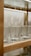A row of textured glass tumblers is displayed on a wooden and glass shelf. The glasses have a diamond or honeycomb pattern and appear to be empty. The shelf is well-lit, highlighting the clarity and design of the glassware.