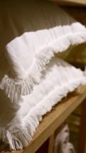 Two white, fringed pillows are stacked neatly on a wooden shelf, creating a cozy and inviting display.