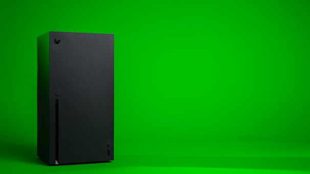 A black, rectangular gaming console stands upright against a vibrant green background. The console has a simple, sleek design with minimalistic features including a disc slot and a few buttons.