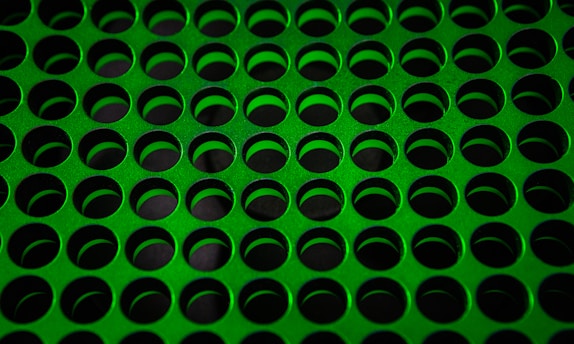 A pattern of evenly spaced, circular holes cut into a green perforated metal sheet, creating a uniform grid across the surface.