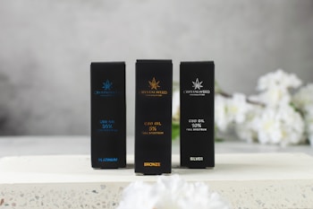 Three rectangular boxes labeled with different types of CBD oil are displayed on a flat surface. The boxes are labeled as Platinum, Bronze, and Silver, each with a different percentage of full-spectrum CBD oil. The background appears to be softly blurred, with white flowers adding a subtle decorative touch.