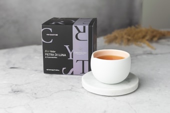A elegant and minimalist setup featuring a cup of tea on a marble coaster, accompanied by a black box labeled 'Pietra di Luna'. The setting is bright with a grey marble surface and a light, neutral background.