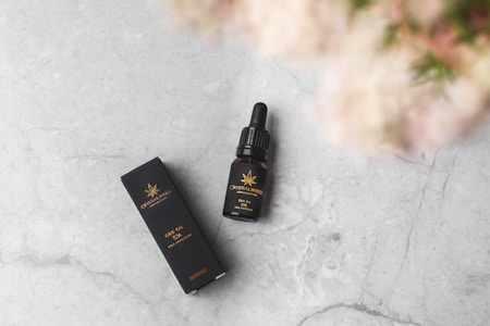 A bottle of CBD oil with a dropper is positioned next to its black packaging box on a light gray marble surface. The product labels are gold and include an emblematic design. In the upper right corner, part of a blurred pink and green floral element is visible.