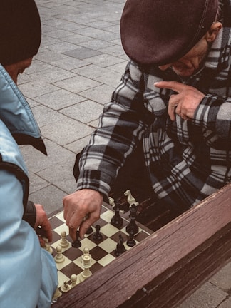 Two individuals are engaged in a game of chess on a tiled outdoor surface. One person is wearing a patterned coat and a flat cap, thoughtfully touching their face while observing the board. The other person's hand is visible, moving a piece. Both individuals seem focused on the game.