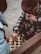 Two individuals are engaged in a game of chess on a tiled outdoor surface. One person is wearing a patterned coat and a flat cap, thoughtfully touching their face while observing the board. The other person's hand is visible, moving a piece. Both individuals seem focused on the game.
