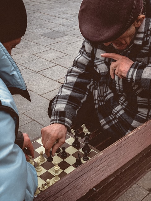 Two individuals are engaged in a game of chess on a tiled outdoor surface. One person is wearing a patterned coat and a flat cap, thoughtfully touching their face while observing the board. The other person's hand is visible, moving a piece. Both individuals seem focused on the game.