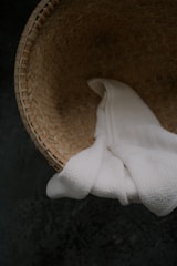 A woven basket with a light brown color pattern contains a soft, white towel placed inside. The basket is situated on a dark textured surface.