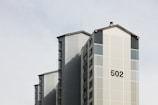 A tall residential building with a minimalist design featuring a prominent number '502' on its facade. The structure includes vertical and horizontal lines, with a light grey color scheme and subtle shadows, set against a pale sky.