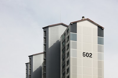 A tall residential building with a minimalist design featuring a prominent number '502' on its facade. The structure includes vertical and horizontal lines, with a light grey color scheme and subtle shadows, set against a pale sky.