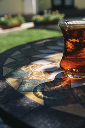A glass of tea sits on a patterned, mosaic-style outdoor table, reflecting sunlight. The background features blurred garden plants and grass, creating a peaceful atmosphere.