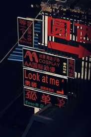 A series of vibrant neon signs with Chinese and English text, featuring phrases like 'Look at me' and mentions of cosplay. The signs are illuminated in bright red and orange, with light reflecting on various surfaces, creating a lively urban atmosphere.