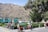 A mountainous landscape in the background with a turquoise building in the middle ground. In the foreground, a woman in traditional clothing holds a colorful alpaca. A statue stands nearby, and there are street vendors with stalls selling various goods under canopies. Trees and people are scattered around the scene.