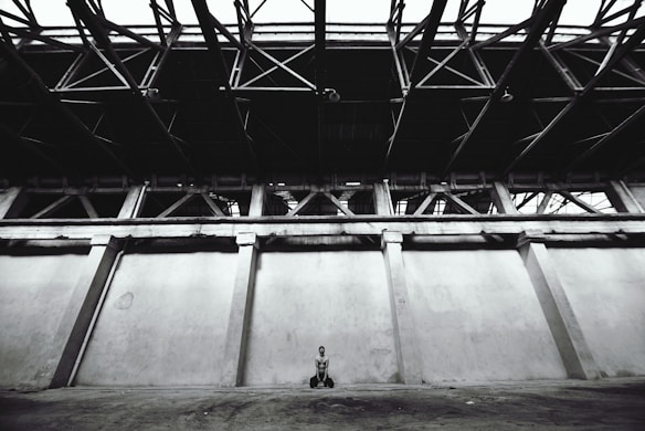 A vast, empty, industrial space with a high ceiling and exposed metal beams. A solitary figure sits on the ground near the center, creating a sense of scale against the large walls and open space.