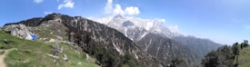 A panoramic view of a mountainous landscape with snow-capped peaks in the distance. The foreground features lush green meadows with scattered rocks and a small tent, indicating a campsite. Dense forested areas cover the slopes, and the sky is clear with a few fluffy clouds.
