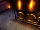 Three large wooden barrels are lined up behind a glass window, illuminated by soft, warm lighting. The floor and surrounding walls are wooden, contributing to a rustic ambiance.