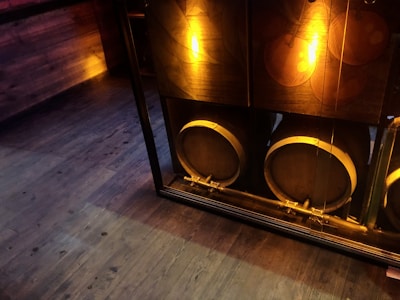 Three large wooden barrels are lined up behind a glass window, illuminated by soft, warm lighting. The floor and surrounding walls are wooden, contributing to a rustic ambiance.