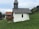 A small white chapel with a wooden shingle roof is situated on a grassy hillside. A cow is standing next to the chapel, partially obscured by the structure. In the background, there are more cows grazing on the grass and another building with a red-tiled roof. The surrounding area features trees and rolling hills under a partly cloudy sky.