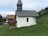 A small white chapel with a wooden shingle roof is situated on a grassy hillside. A cow is standing next to the chapel, partially obscured by the structure. In the background, there are more cows grazing on the grass and another building with a red-tiled roof. The surrounding area features trees and rolling hills under a partly cloudy sky.
