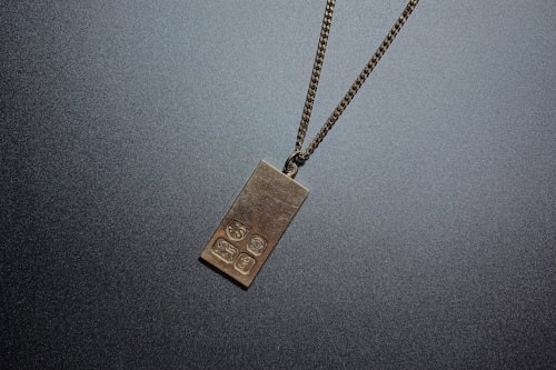 A metal pendant in the shape of a rectangular gold bar hangs from a fine chain. The pendant has several engraved symbols on its surface.