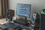A modern desktop setup featuring a monitor displaying financial market charts, accompanied by a keyboard, mouse, and stylish speakers. A stack of notebooks with a pen rests beside a motivational coffee cup. A small calendar displaying the year 2021 is present, and a pair of headphones hang on a speaker.