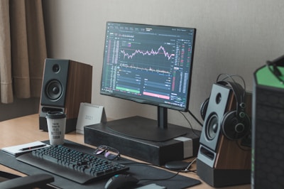 A modern desktop setup featuring a monitor displaying financial market charts, accompanied by a keyboard, mouse, and stylish speakers. A stack of notebooks with a pen rests beside a motivational coffee cup. A small calendar displaying the year 2021 is present, and a pair of headphones hang on a speaker.