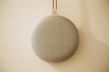 A circular, perforated speaker hangs against a light-colored wall, creating a minimalist and modern aesthetic. The speaker's surface features a grid of small holes, and it is suspended by a thin cord with a small logo at the top.