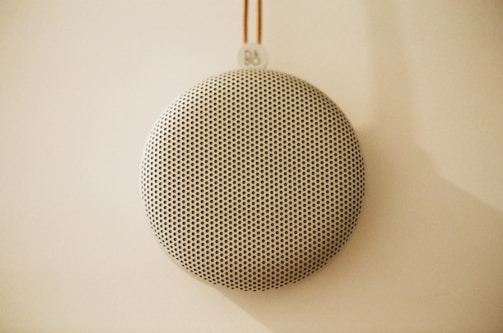 A circular, perforated speaker hangs against a light-colored wall, creating a minimalist and modern aesthetic. The speaker's surface features a grid of small holes, and it is suspended by a thin cord with a small logo at the top.