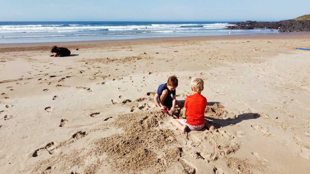 2 boys sitting on brown sand near sea during daytime
