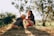 A young woman is kneeling on the grass, gently touching a golden retriever wearing a shirt. They are surrounded by greenery and trees, and the sunlight creates a warm and peaceful atmosphere.