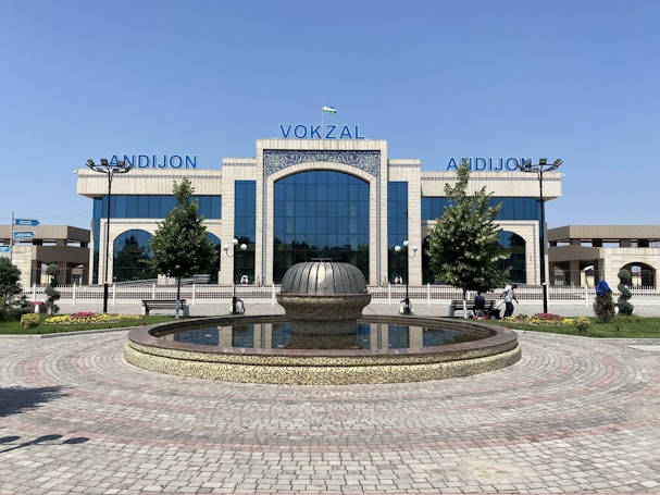 A modern building with large glass windows and the word 'VOKZAL' prominently displayed in blue above the entrance. In the foreground, a circular fountain with decorative stonework is surrounded by neatly arranged paving stones. There are small trees and landscaped plants on either side of the entrance, and a few people are seated on benches or walking nearby.