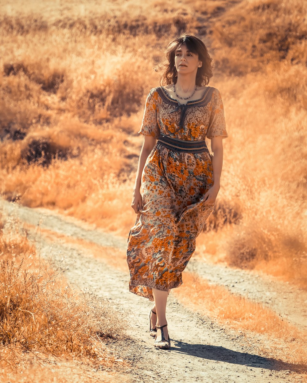 Mujer en vestido floral marrón y negro de pie en el camino de tierra durante el día