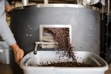 A large coffee roasting machine releases a stream of freshly roasted coffee beans into a nearby tray. A person is seen handling the equipment while beans cascade down. The setting appears to be a coffee roastery or industrial workspace.